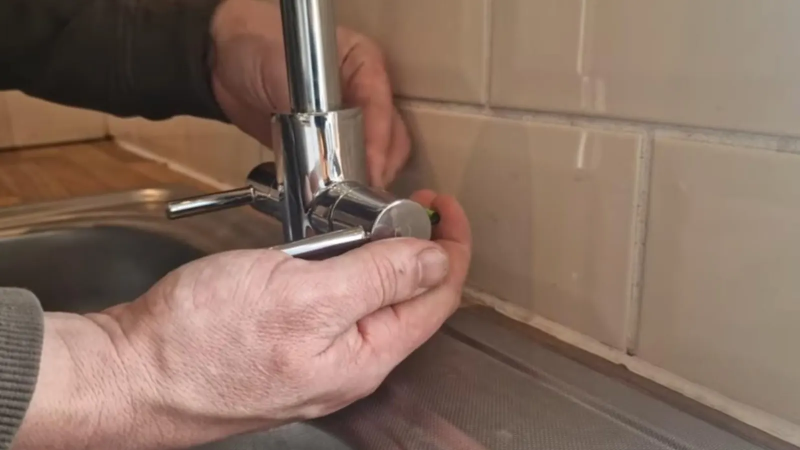 Hands installing a chrome kitchen faucet at a sink with tiled backsplash