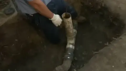 Plumber kneeling in an excavated trench working on a buried sewer pipe